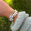 A close-up of a person's wrist wearing a stack of beaded bracelets, including white pearl-like beads, light blue beads, olive green beads, and small gold accents, with a light blue knit sleeve visible against a blurred green outdoor background.