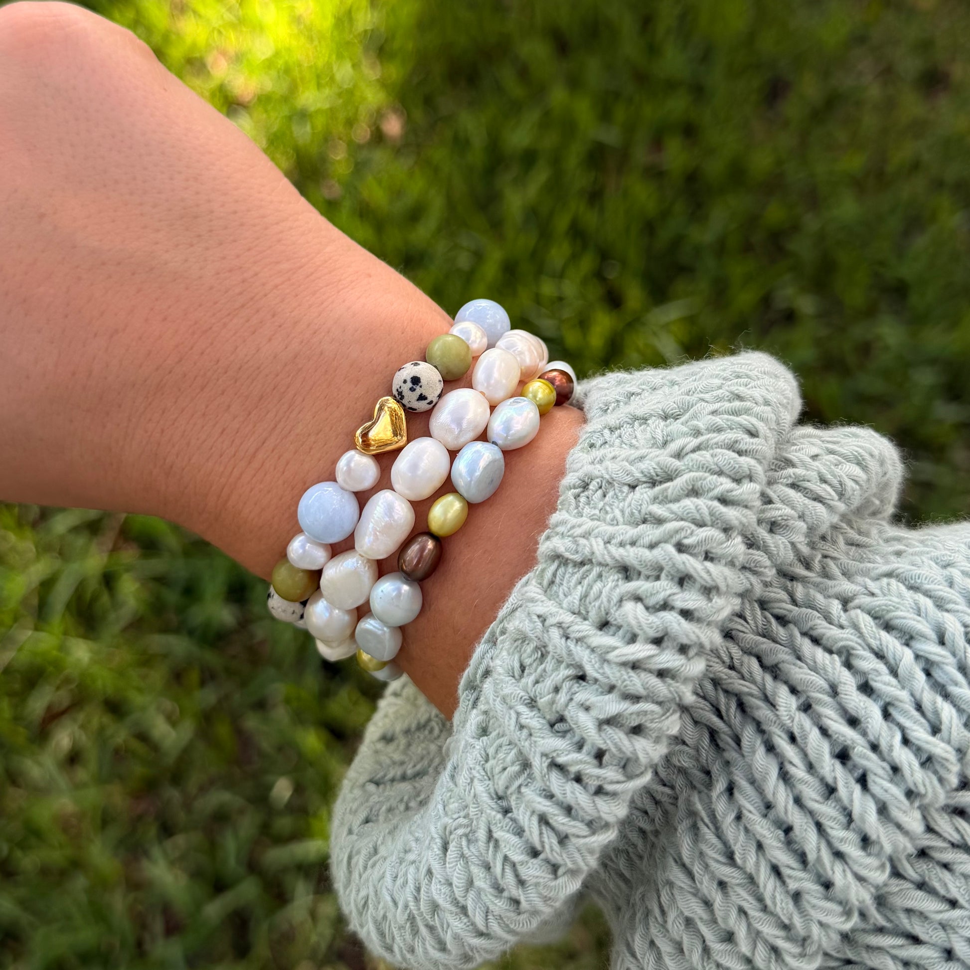 A close-up of a person's wrist wearing a stack of beaded bracelets, including white pearl-like beads, light blue beads, olive green beads, and small gold accents, with a light blue knit sleeve visible against a blurred green outdoor background.