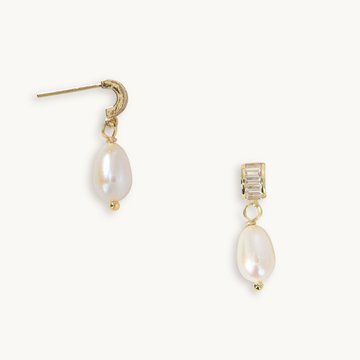 A pair of gold drop earrings featuring a textured gold crescent-shaped stud on one earring and a gold rectangular stud with clear baguette-cut crystals on the other, both hanging from gold posts with teardrop-shaped white freshwater pearls below.