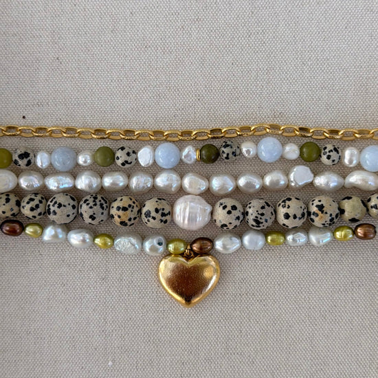 A close-up of a multi-strand beaded bracelet laid flat on a neutral beige fabric background. The bracelet features a gold-tone chain, white baroque pearls, speckled beige and black beads, olive green beads, and a shiny gold heart charm.