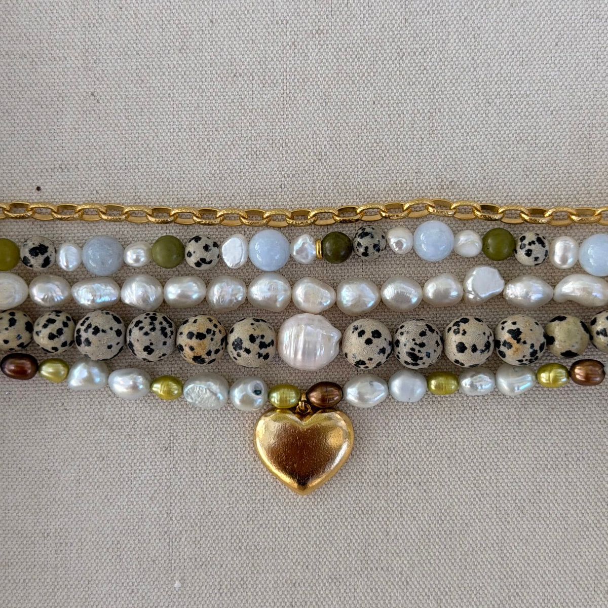 A close-up of a multi-strand beaded bracelet laid flat on a neutral beige fabric background. The bracelet features a gold-tone chain, white baroque pearls, speckled beige and black beads, olive green beads, and a shiny gold heart charm.