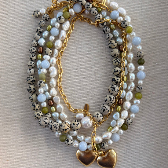A multi-strand beaded necklace with white pearls, black-and-white spotted beads, light blue beads, olive green beads, and gold-tone chains with two gold heart charms, displayed on a neutral fabric background.