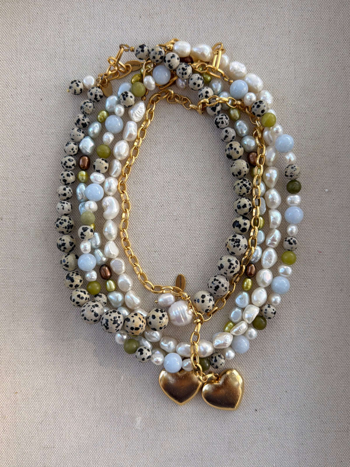 A multi-strand beaded necklace with white pearls, black-and-white spotted beads, light blue beads, olive green beads, and gold-tone chains with two gold heart charms, displayed on a neutral fabric background.