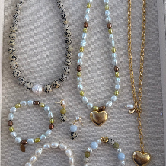A jewelry set displayed on a beige fabric-lined tray, featuring multiple beaded necklaces, bracelets, earrings, and a gold heart pendant necklace.