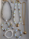 A jewelry set displayed on a beige fabric-lined tray, featuring multiple beaded necklaces, bracelets, earrings, and a gold heart pendant necklace.