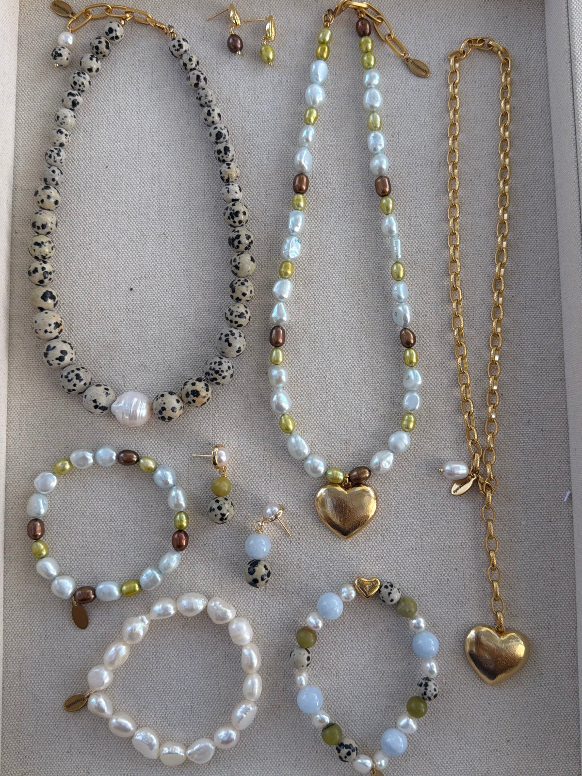 A jewelry set displayed on a beige fabric-lined tray, featuring multiple beaded necklaces, bracelets, earrings, and a gold heart pendant necklace.