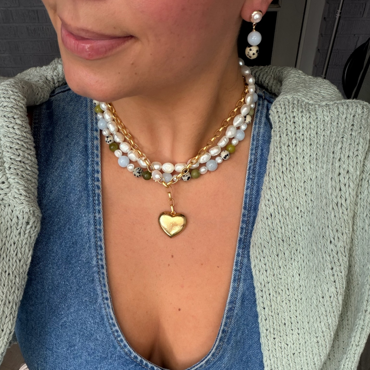 A close-up of a person wearing a layered necklace with a gold heart pendant, mixed white and green beads, and gold chain links, styled over a denim top and light green knit cardigan.