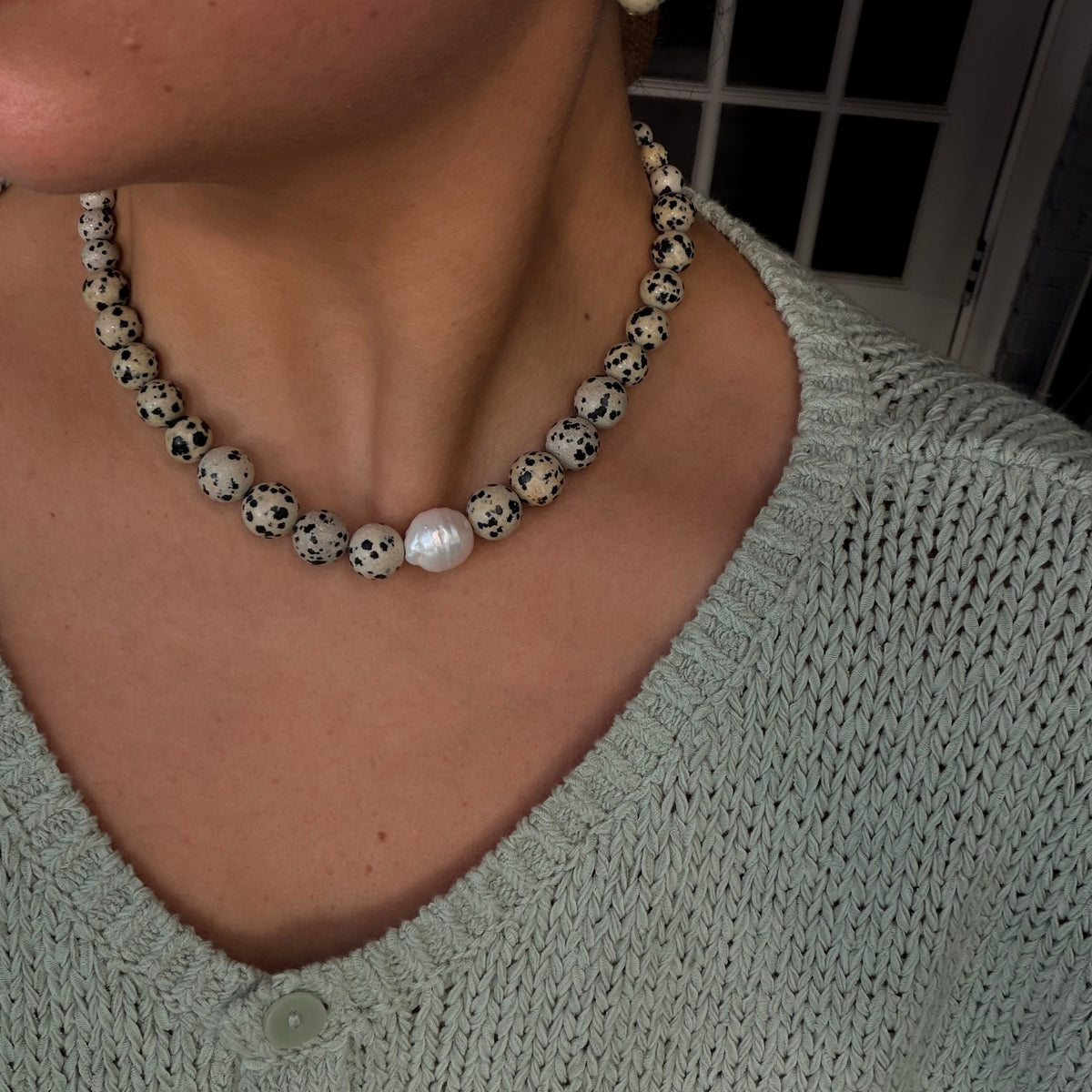 A close-up of a person wearing a Jasper beaded pearl necklace and matching drop earrings. The necklace features speckled white and black beads with a single white pearl accent at the center, worn over a light sage green knit top.