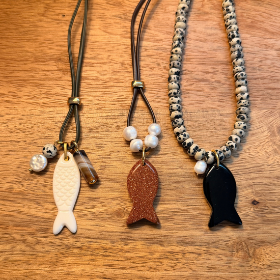 Three fish-shaped pendant necklaces displayed on a wooden surface. The left necklace has a dark cord, a white fish pendant, and small pearl-like beads.
