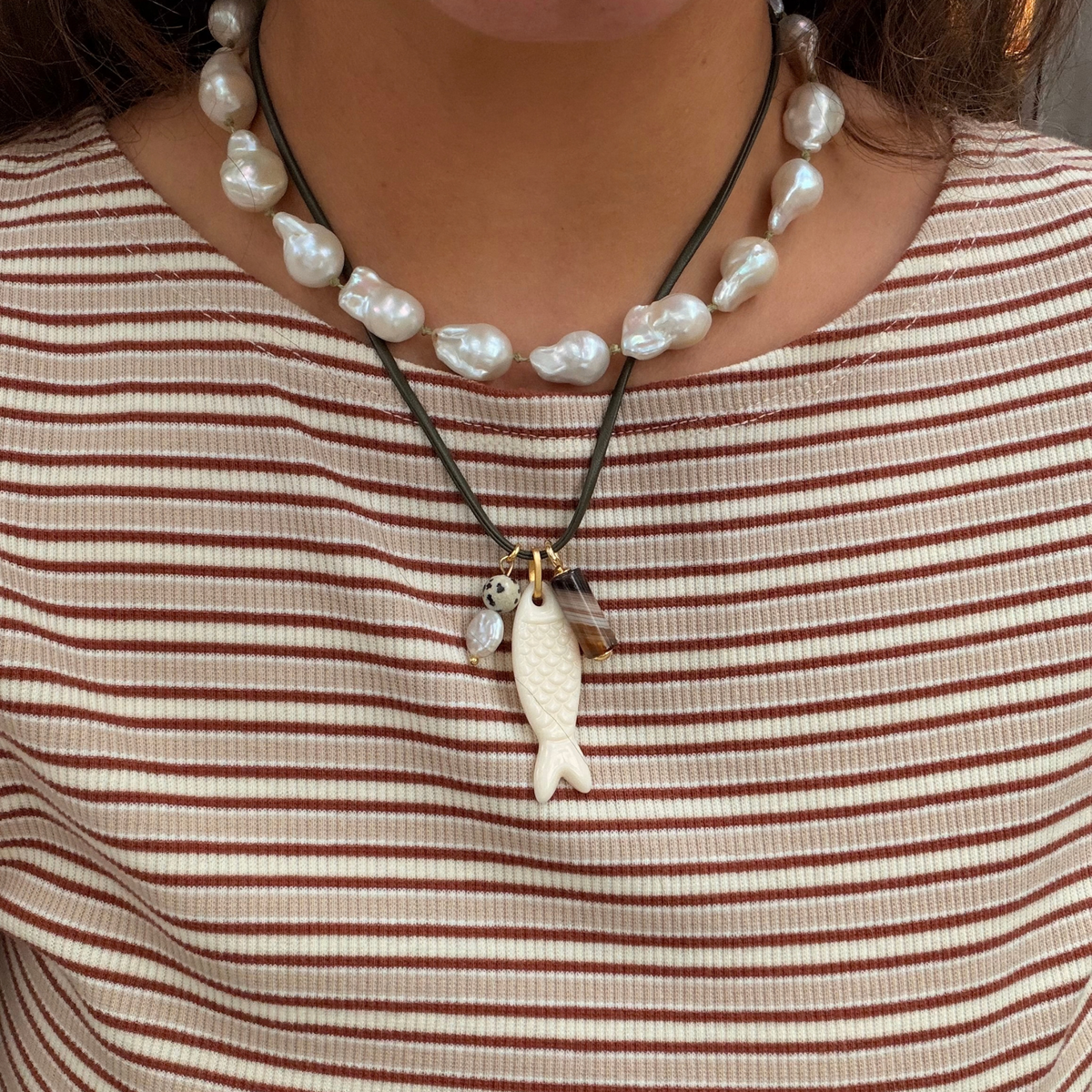 A close-up of a person wearing a layered necklace set. The top necklace features irregular white baroque pearls on a dark cord, while the shorter necklace below has a dark cord with a cream-colored fish pendant and small gold-toned charms.