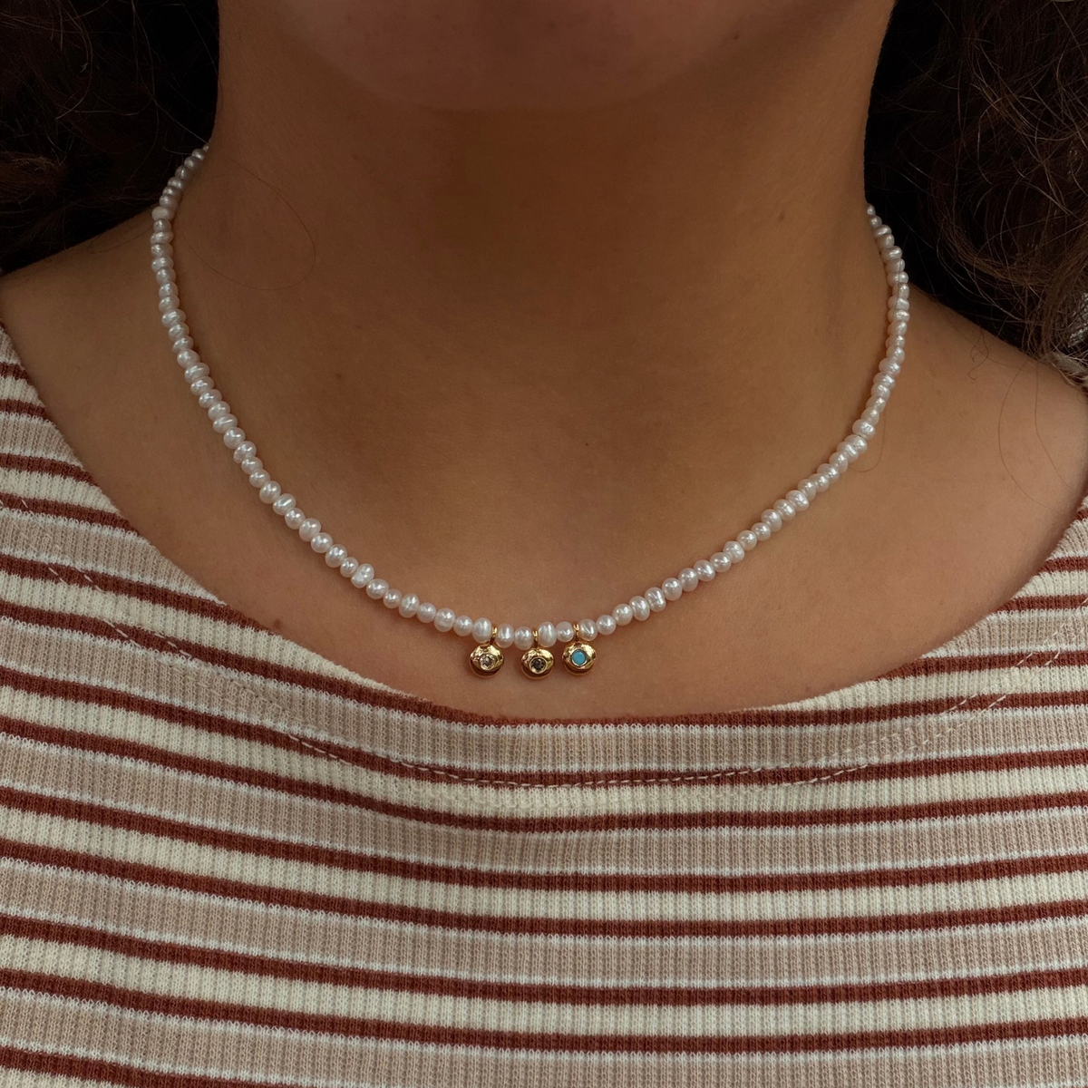 A close-up of a person wearing a delicate pearl necklace with three small gold charms at the center, including one with a tiny blue stone, paired with a beige and brown striped top.