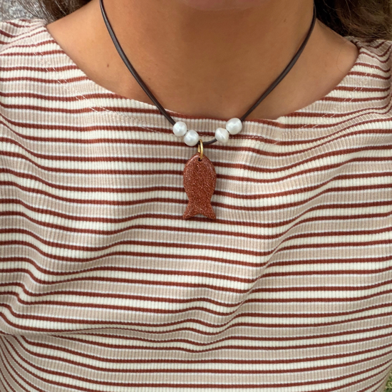 A close-up of a person wearing a necklace with a brown leather cord, white pearl-like beads, and a textured reddish-brown fish-shaped pendant. The person is wearing a beige and brown horizontally striped ribbed top.