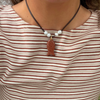 A close-up of a person wearing a necklace with a brown leather cord, white pearl-like beads, and a textured reddish-brown fish-shaped pendant. The person is wearing a beige and brown horizontally striped ribbed top.
