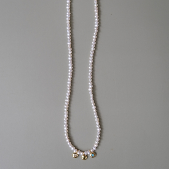 A long strand of white freshwater pearls draped vertically on a neutral gray background, featuring three small gold-toned decorative charms at the bottom.