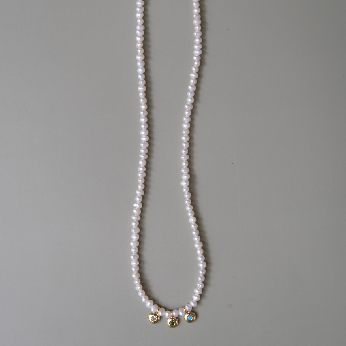 A long strand of white freshwater pearls draped vertically on a neutral gray background, featuring three small gold-toned decorative charms at the bottom.