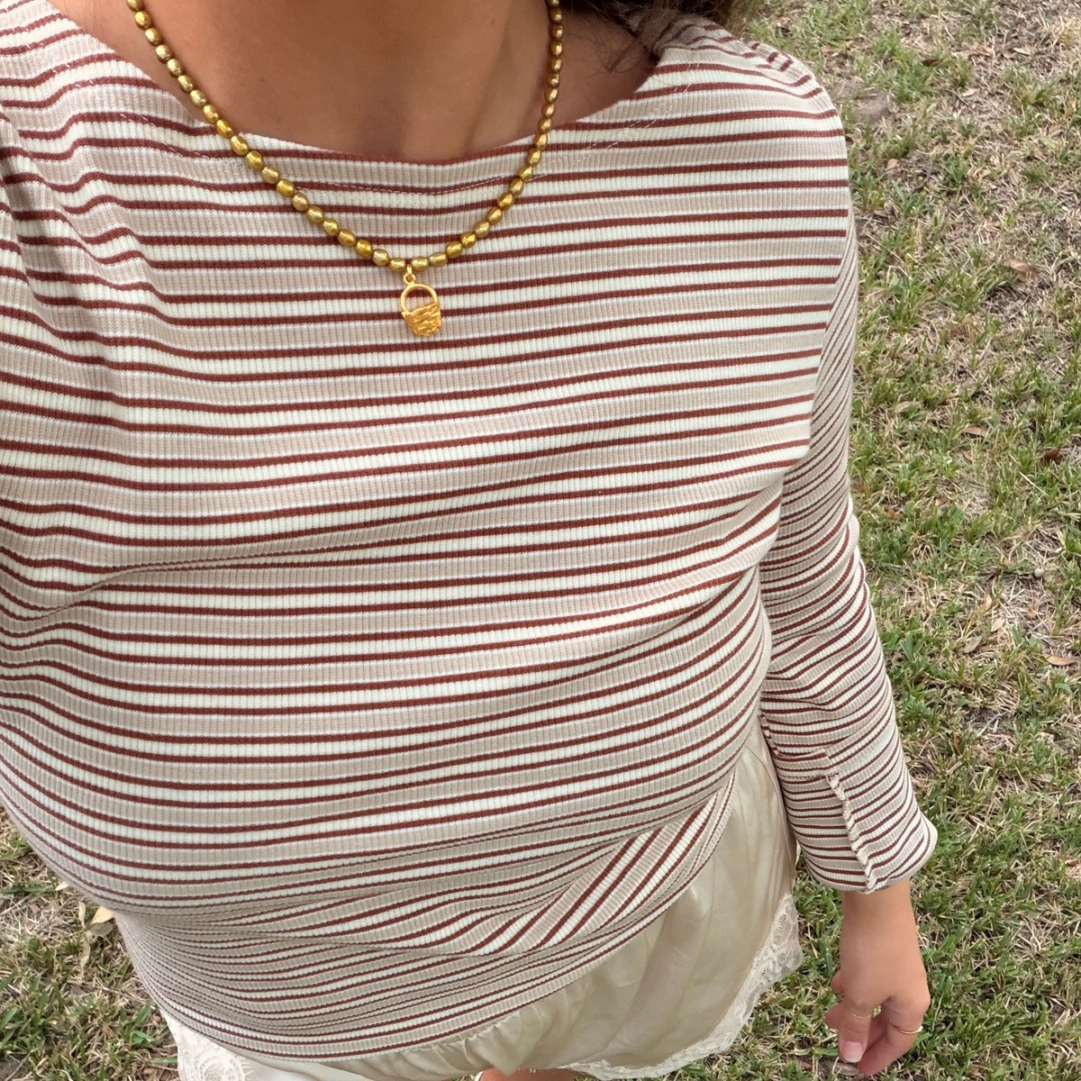 A close-up of a person wearing a beige and brown horizontally striped long-sleeve top, paired with a golden green pearl necklace, standing outdoors on grass.