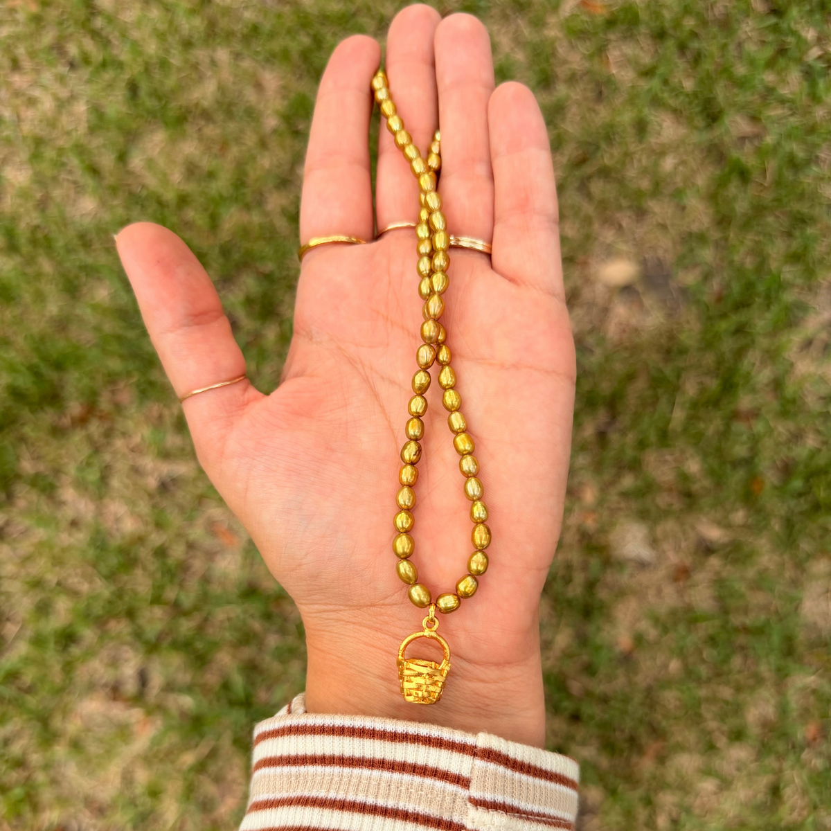 A golden green pearl necklace with a small basket pendant is held in a person's open hand, with gold rings on the fingers, against a blurred green grass background.
