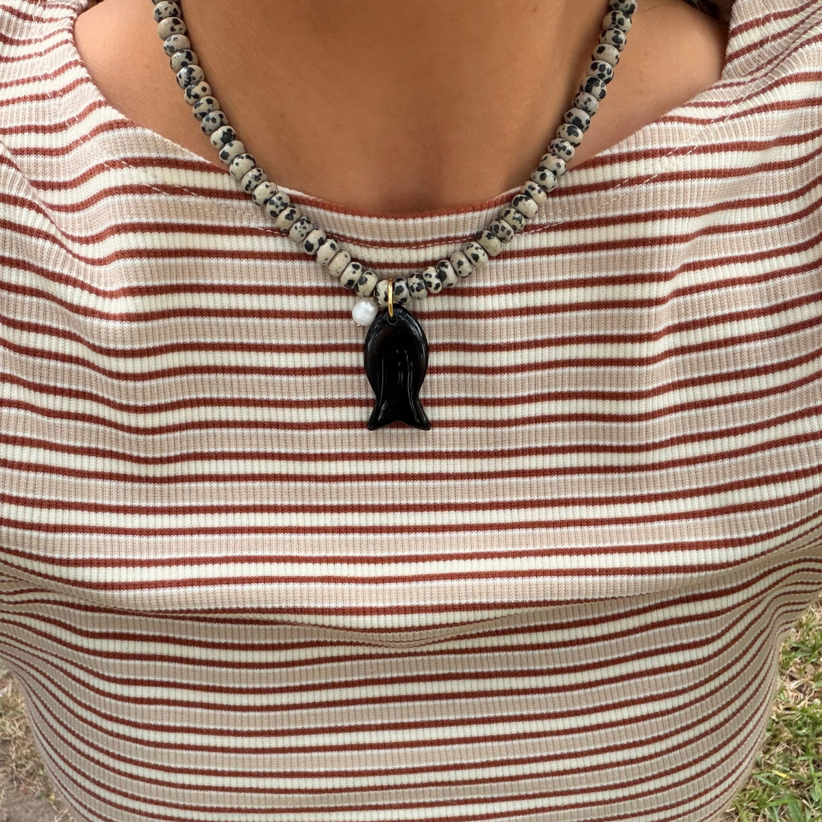 A close-up of a person wearing a beaded necklace with a black pendant over a beige and brown horizontally striped shirt.
