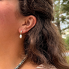 A close-up of a person's ear wearing a gold hoop earring with a small clear stone and a dangling white pearl bead, shown outdoors with blurred green foliage in the background.