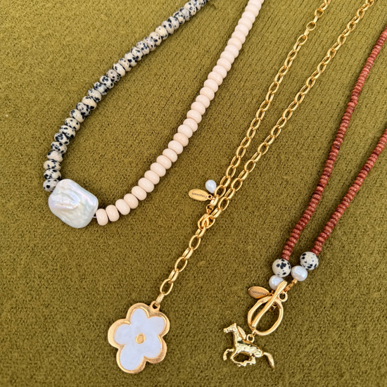 Three handcrafted necklaces on an olive green knit. A strand of dalmatian-spotted and cream rondelles centers on a large baroque pearl, beside a golden chain with a white flower pendant and a rust-red strand finished with a golden horse charm.