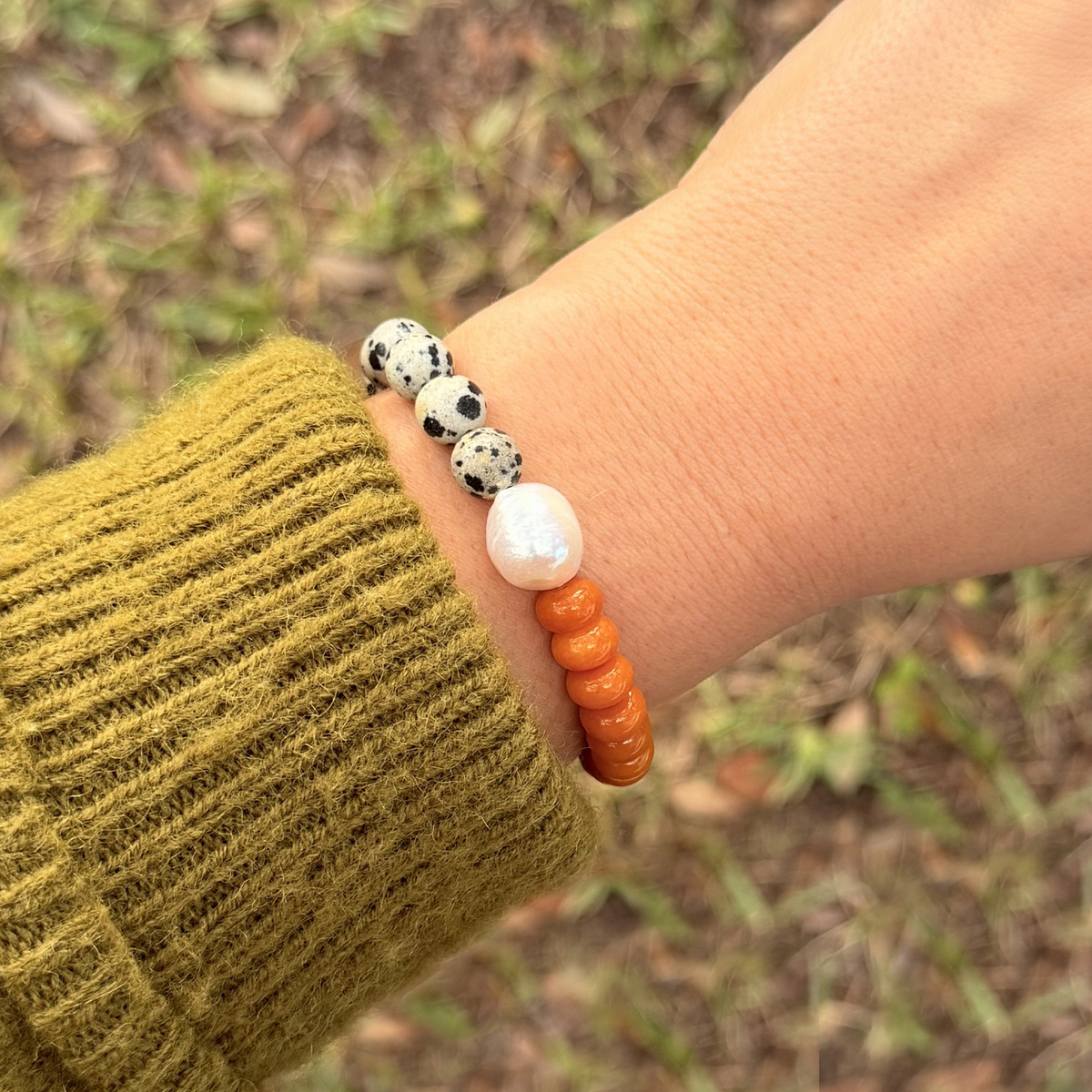 Wrist in an olive sweater wearing the dalmatian and orange bracelet with its freshwater pearl centerpiece, against grass and fallen leaves.