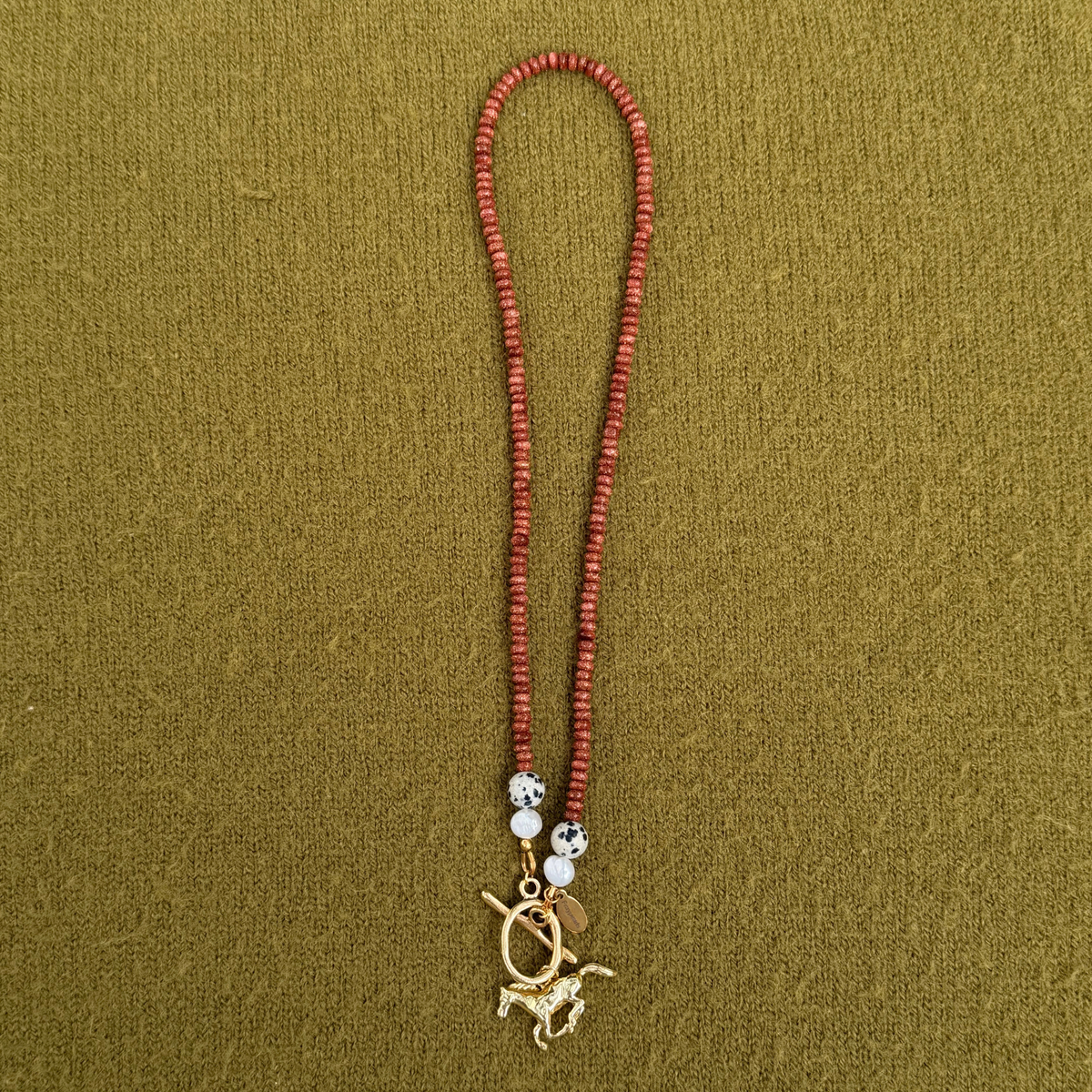 Rust-red sparkly rondelle necklace with dalmatian stones and pearls at each end, joined by a golden toggle clasp with a galloping horse charm.