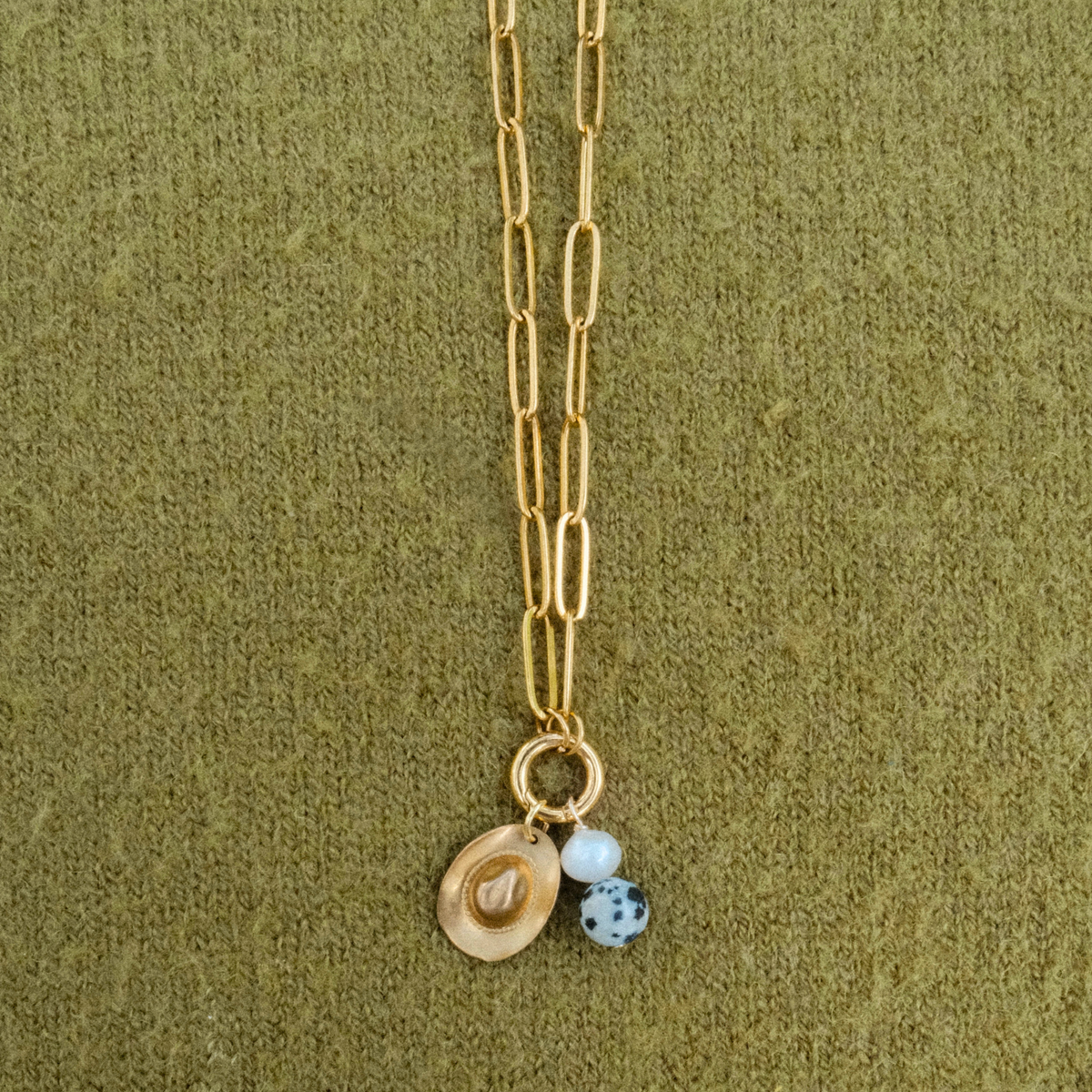 Close-up of a golden paperclip chain with a cowboy hat charm, a freshwater pearl and a dalmatian-spotted stone hanging from a round clasp.