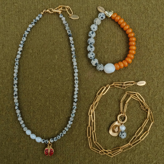Flat lay of the dalmatian ladybug necklace with its pearl section, the dalmatian and orange heart bracelet, and a coiled golden paperclip chain with a cowboy hat charm.