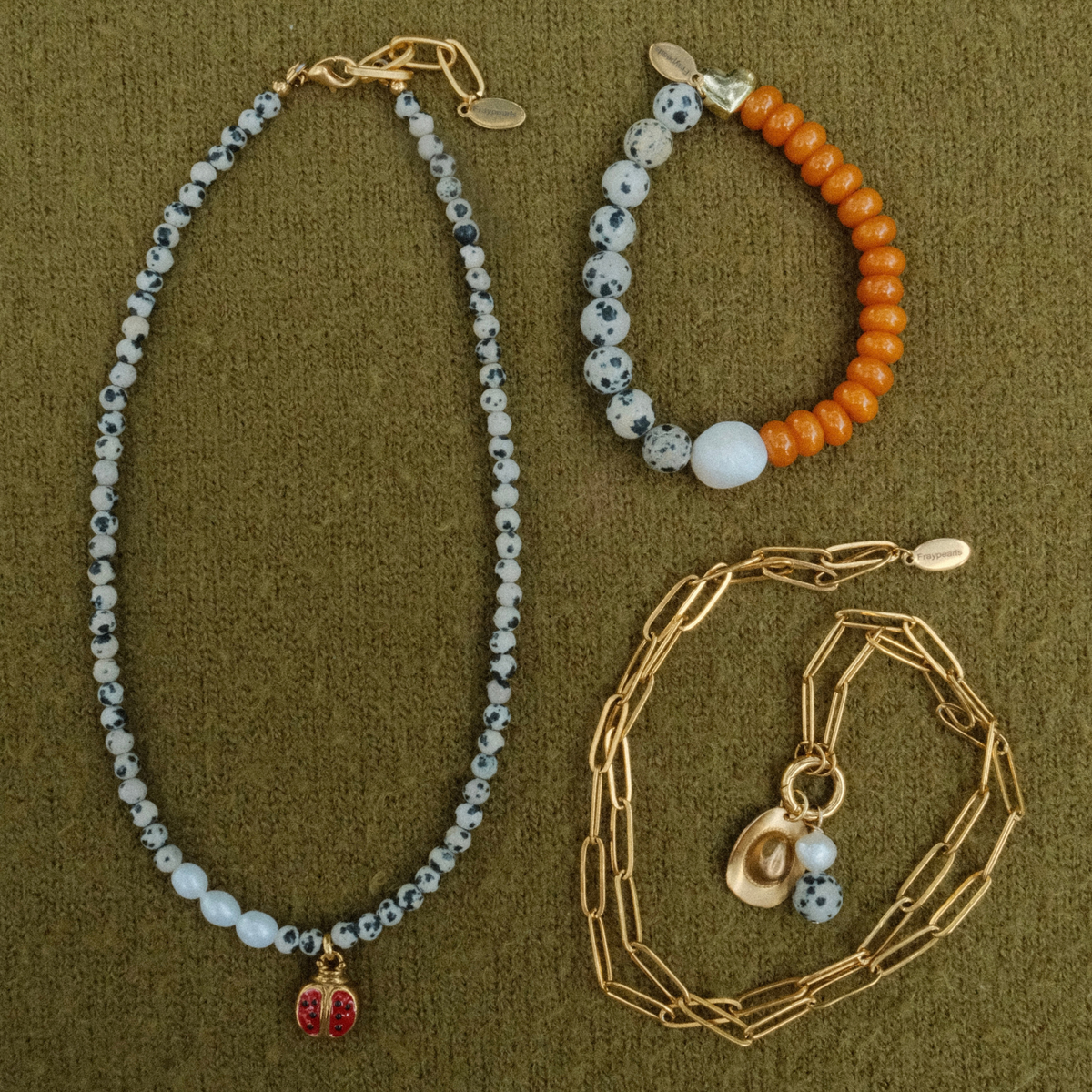 Flat lay of the dalmatian ladybug necklace with its pearl section, the dalmatian and orange heart bracelet, and a coiled golden paperclip chain with a cowboy hat charm.