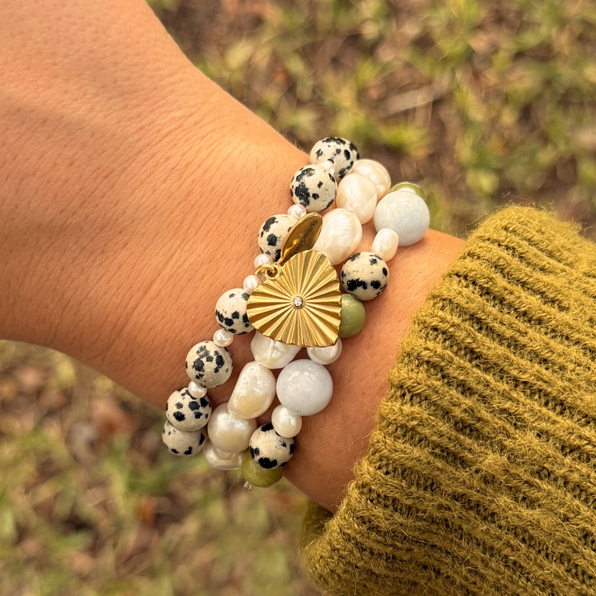 Wrist stacked with the dalmatian and pearl heart bracelet and a second bracelet of baroque pearls with pale blue and green stones, against grass.