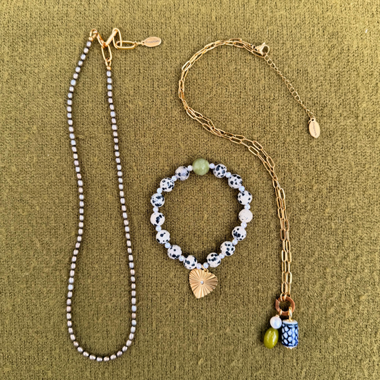 Flat lay of a slim strand of gray freshwater pearls, a dalmatian and pearl bracelet with a golden heart charm, and a golden paperclip chain holding a pearl, green stone and blue and white ceramic charm.