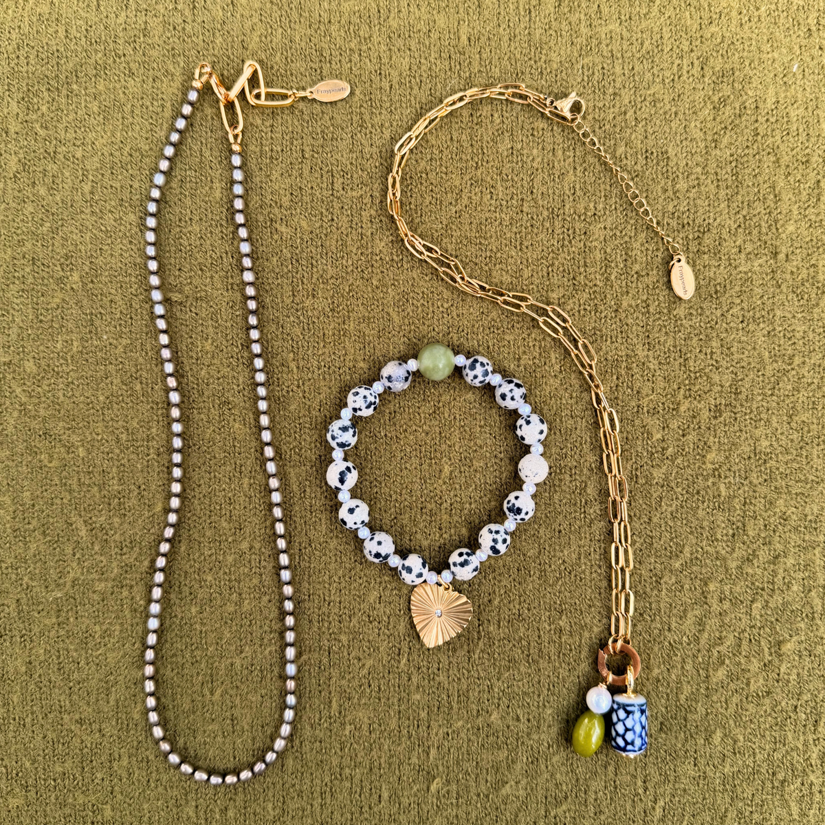 Flat lay of a slim strand of gray freshwater pearls, a dalmatian and pearl bracelet with a golden heart charm, and a golden paperclip chain holding a pearl, green stone and blue and white ceramic charm.