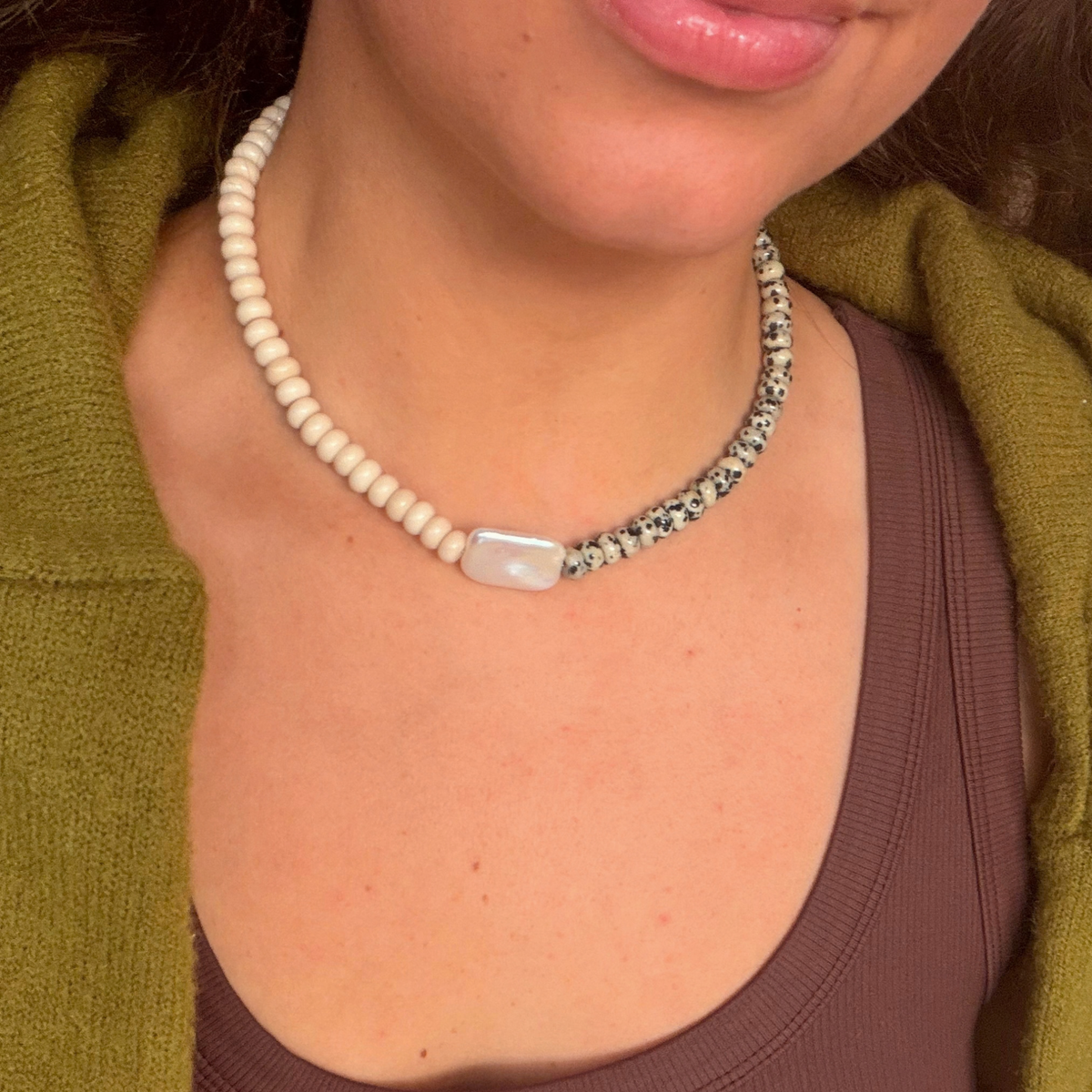 Close-up of a strand of cream rondelles and dalmatian-spotted stones meeting at a rectangular baroque pearl, worn with a brown tank and olive cardigan.