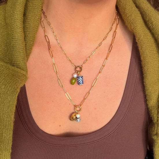 Two layered golden paperclip chains. The shorter holds a pearl, green stone and blue and white ceramic charm, and the longer holds a golden cowboy hat charm, a pearl and a dalmatian stone.
