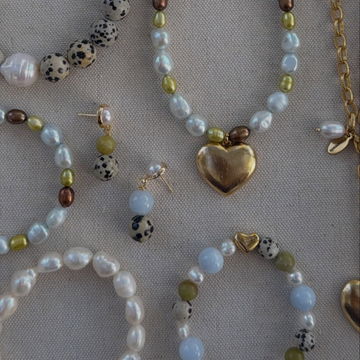 A styled flat-lay of mixed beaded jewelry on a textured beige fabric background, featuring a gold heart pendant necklace, matching earrings, bracelets, and beaded strands with pearls, olive green, brown, and spotted beads.