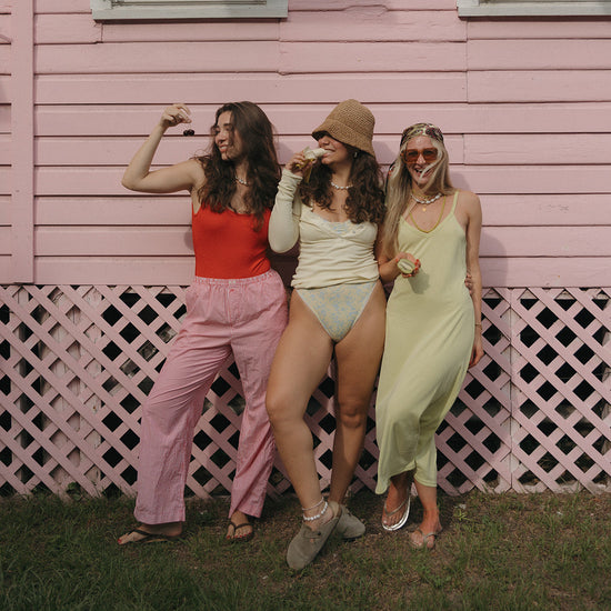 Three women smiling in front of a pink wooden wall, each holding fruits. They wear colorful summer outfits with pearl jewelry, exuding a cheerful, relaxed vibe.