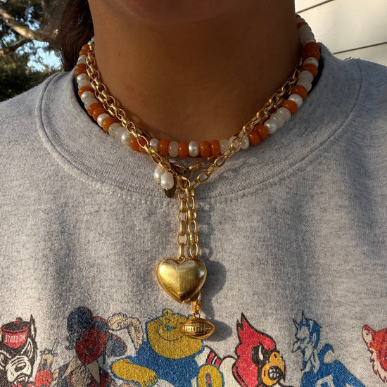 A close-up of a person wearing a layered necklace with gold chain, white and orange beads, and a gold heart pendant, paired with a gray graphic sweatshirt.