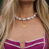 Close-up of a woman's neck adorned with a chunky pearl necklace and a gold pendant necklace. She wears a magenta top with white trim.
