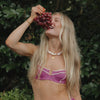 A woman with long blonde hair joyfully holds a bunch of red grapes near her mouth. She is outdoors, wearing a pink bikini top and a pearl necklace, surrounded by lush greenery.