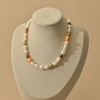 A necklace displayed on a neutral beige jewelry bust against a warm beige background. The necklace features irregular white baroque pearls interspersed with small orange, green, and brown peals.