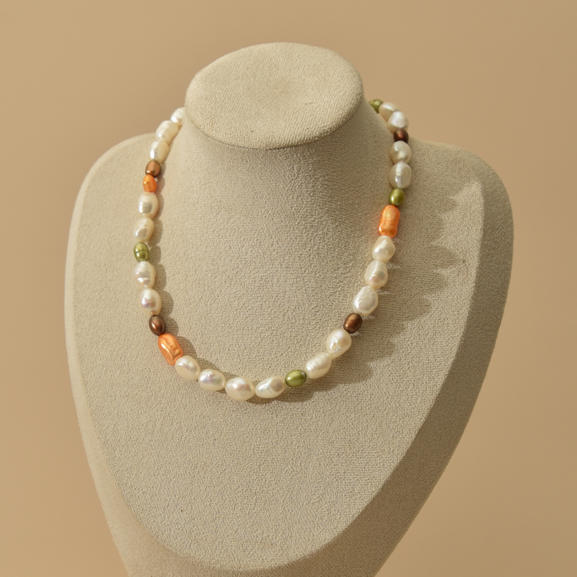 A necklace displayed on a neutral beige jewelry bust against a warm beige background. The necklace features irregular white baroque pearls interspersed with small orange, green, and brown peals.