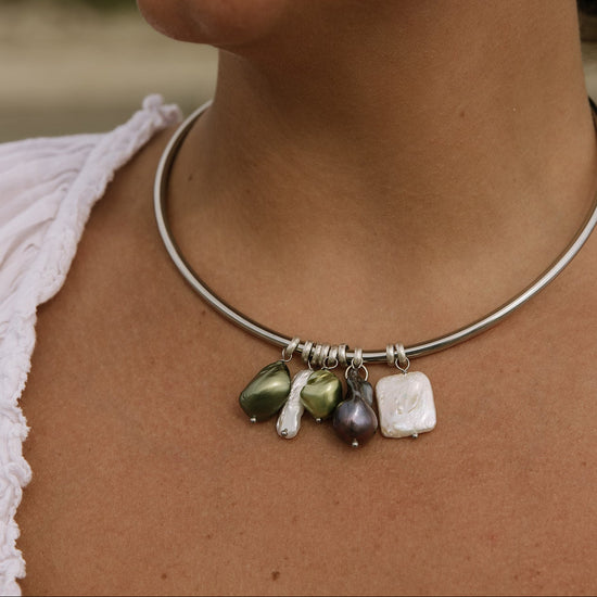 A close-up of a person wearing a thin silver-toned bangle necklace with dangling baroque-style beads in olive green, white, and dark purple.