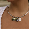 A close-up of a person wearing a thin silver-toned bangle necklace with dangling baroque-style beads in olive green, white, and dark purple.