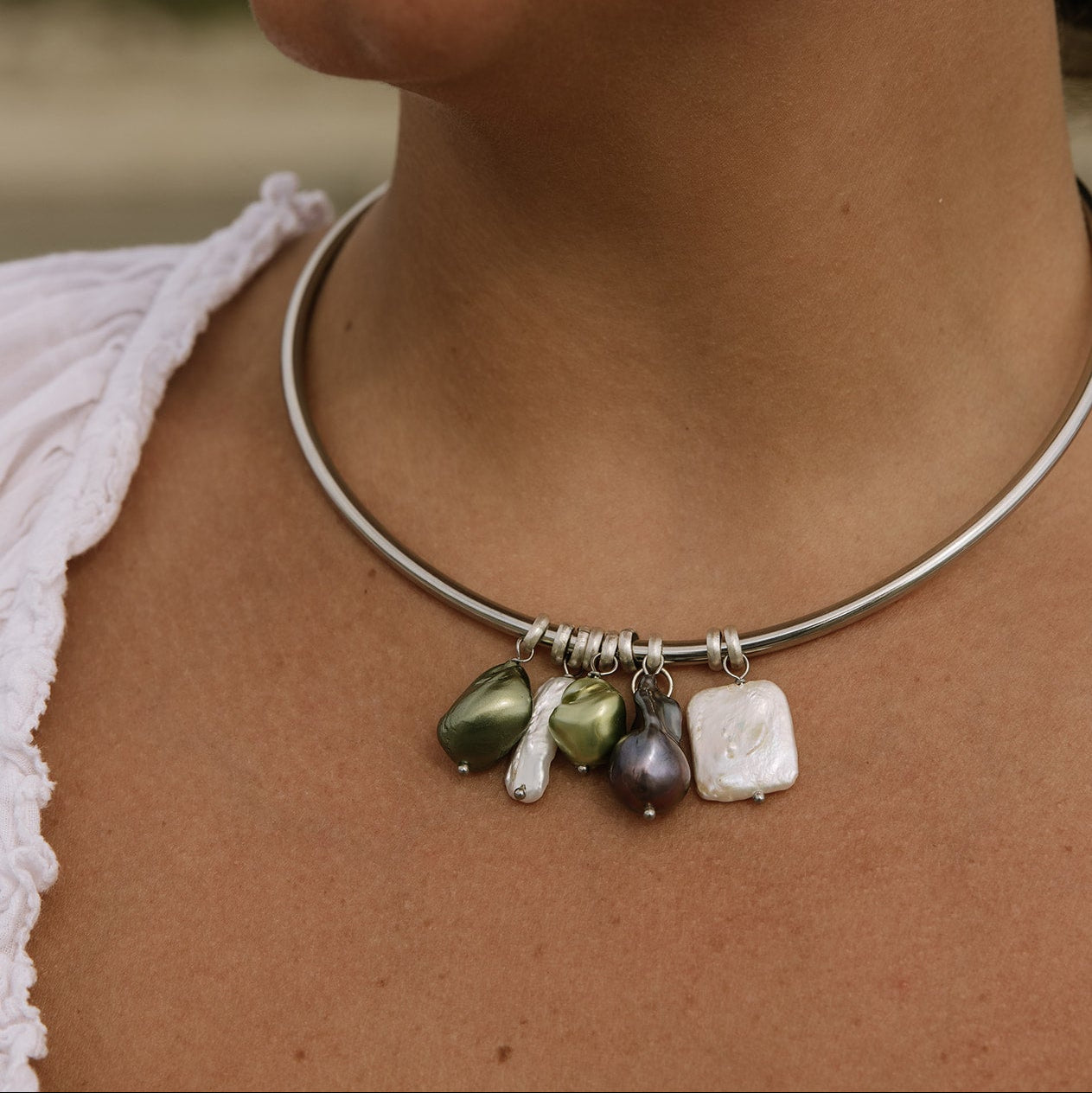 A close-up of a person wearing a thin silver-toned bangle necklace with dangling baroque-style beads in olive green, white, and dark purple.