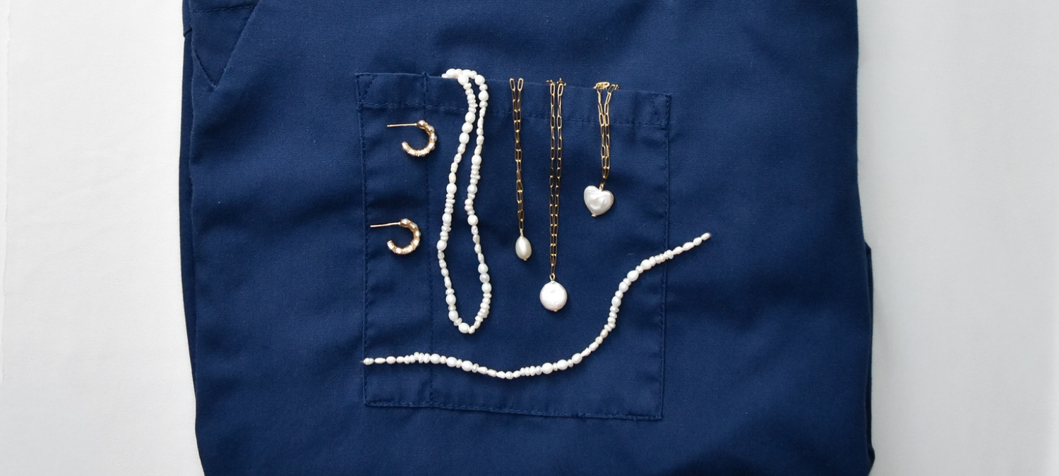 Folded navy blue scrub top adorned with gold and pearl necklaces draped over the front pocket, against a plain white background.