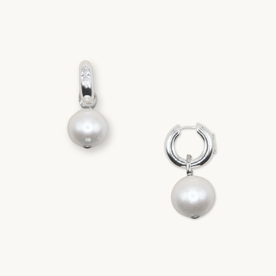 Silver hoop earrings each featuring a dangling white pearl. One earring shows a small, clear gemstone on the hoop. Set against a plain background.