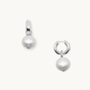 Silver hoop earrings each featuring a dangling white pearl. One earring shows a small, clear gemstone on the hoop. Set against a plain background.