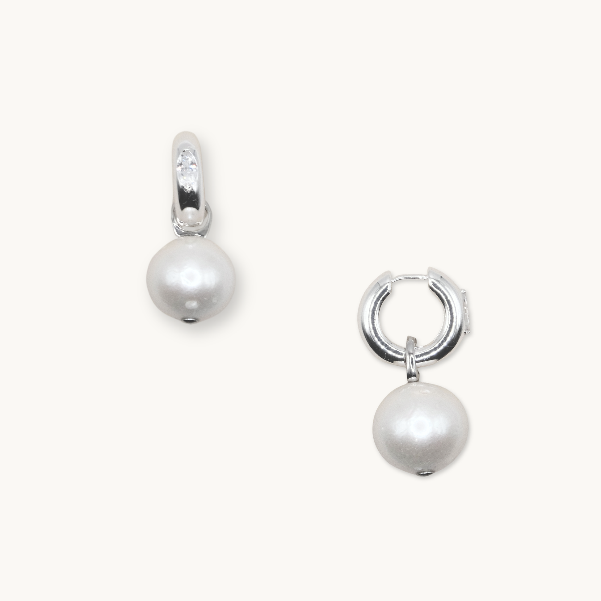 Silver hoop earrings each featuring a dangling white pearl. One earring shows a small, clear gemstone on the hoop. Set against a plain background.