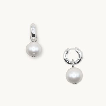 Silver hoop earrings each featuring a dangling white pearl. One earring shows a small, clear gemstone on the hoop. Set against a plain background.
