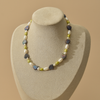 A pearl necklace displayed on a beige jewelry bust against a warm beige background. The necklace features irregular dark blue pearls, white baroque pearls, and small green pearls.